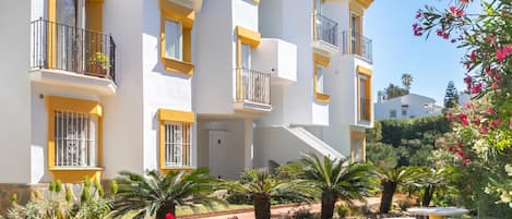 A multi-story building with balconies, a garden with palm trees, and a stone pathway.