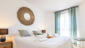 A neatly made bed with pillows, a bedside table with a lamp, a decorative mirror, and a window with curtains. - Calahonda Golf (Mijas)