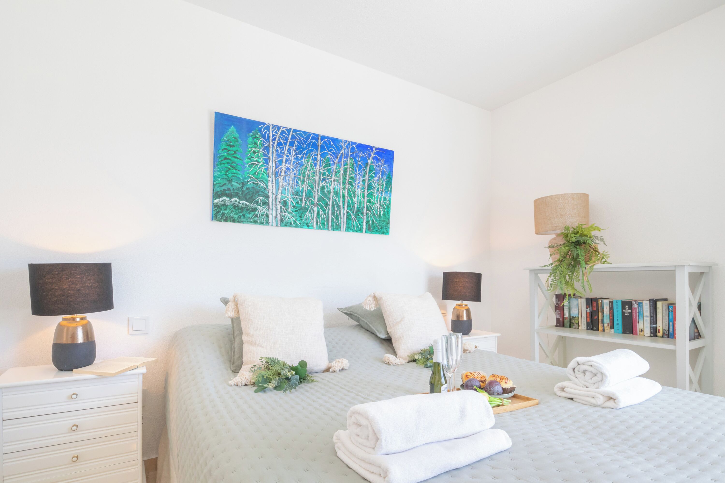 A neatly arranged bedroom with a bed, bedside tables, a bookshelf, and a painting on the wall.