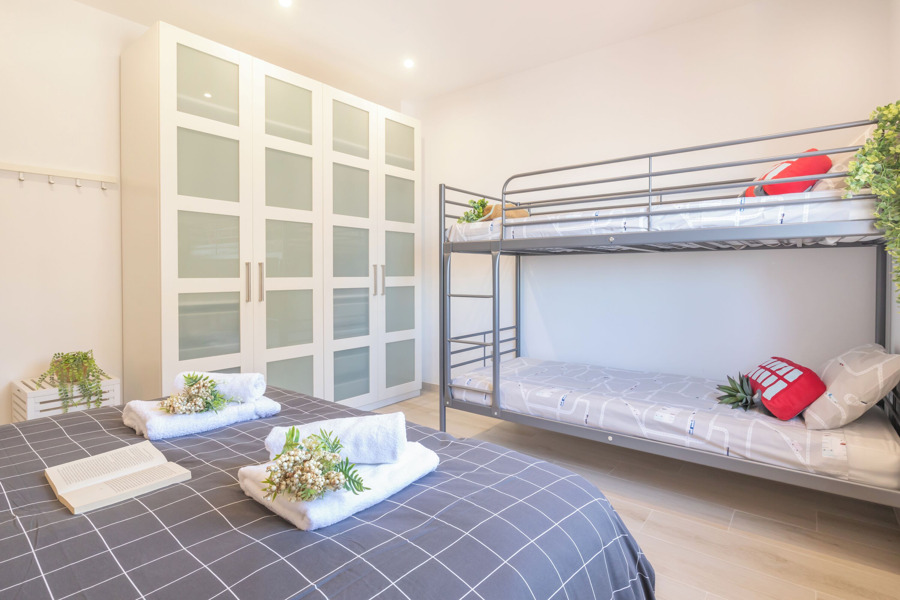Une chambre avec un lit superposé, une grande armoire aux portes vitrées, un lit recouvert d’une couverture à motifs sombres et une petite plante posée sur le sol.
