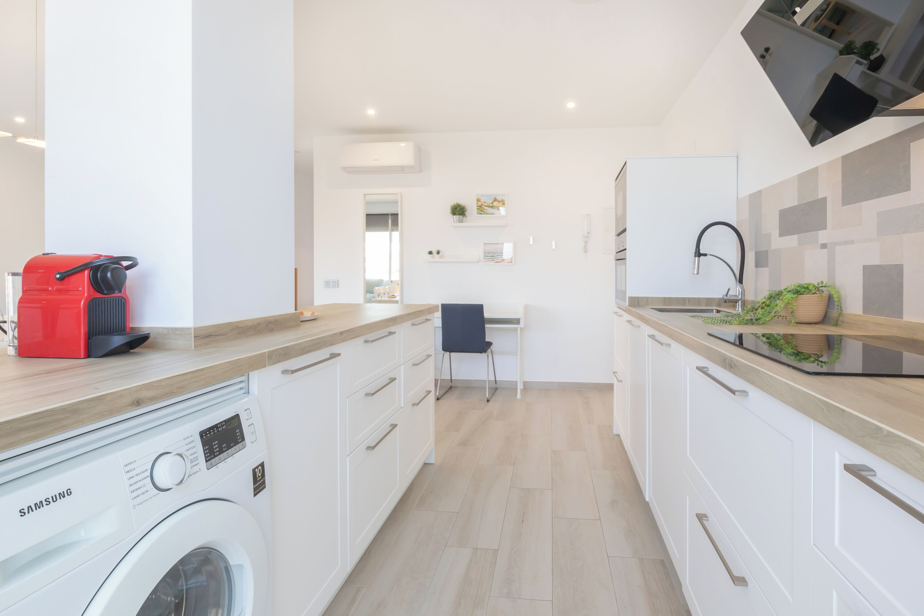 A modern kitchen with a washing machine, a red coffee machine, and a dining area with a chair and table.