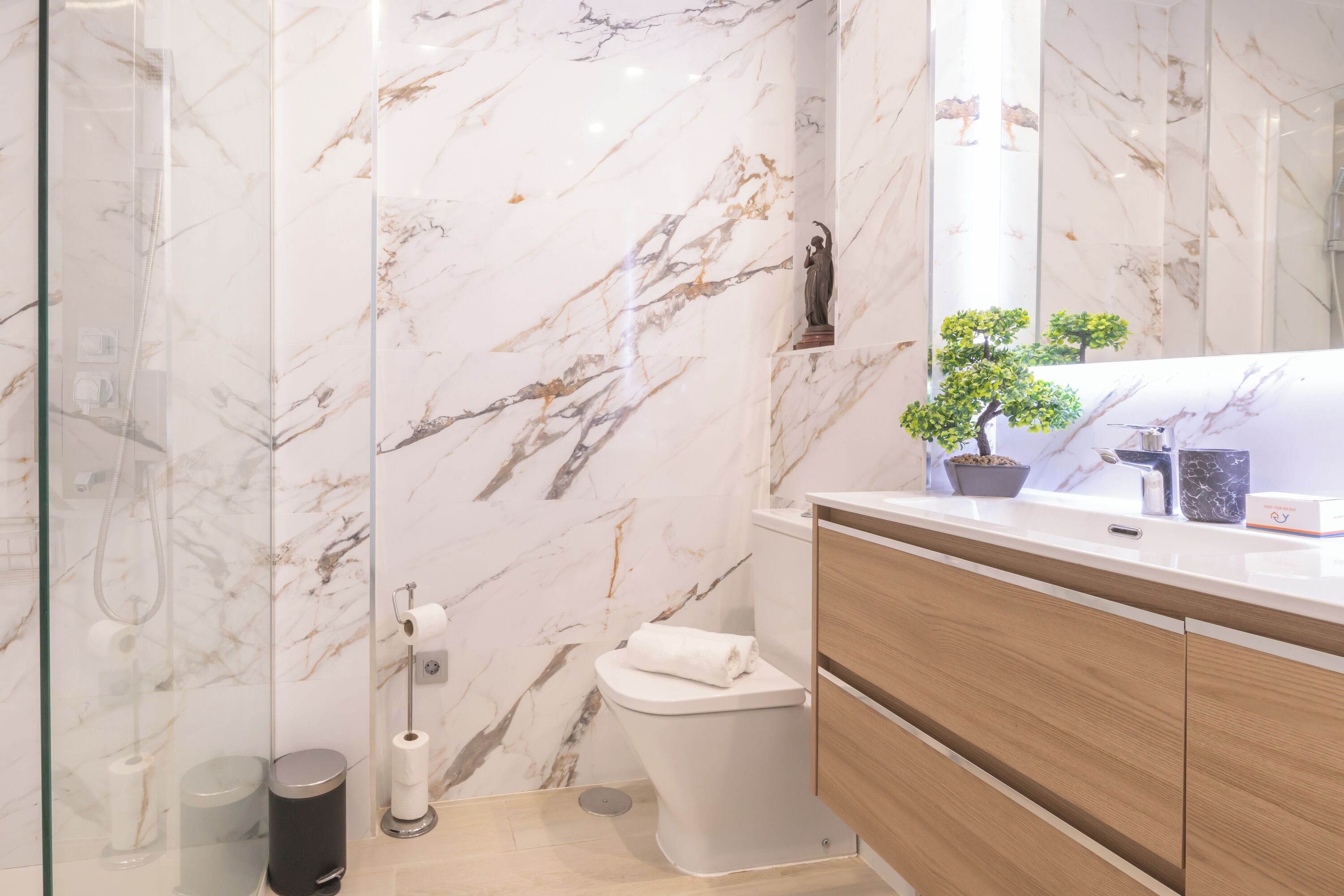 A modern bathroom with marble walls, a glass shower enclosure, a toilet, and a wooden vanity with a mirror.