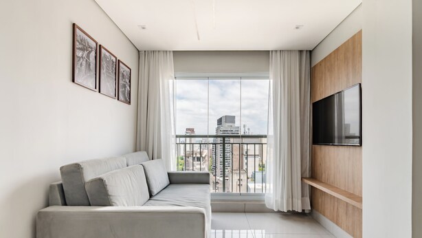 A modern living room with a grey sofa, a mounted TV, and a balcony with a city view. - 360 Paraiso (São Paulo)