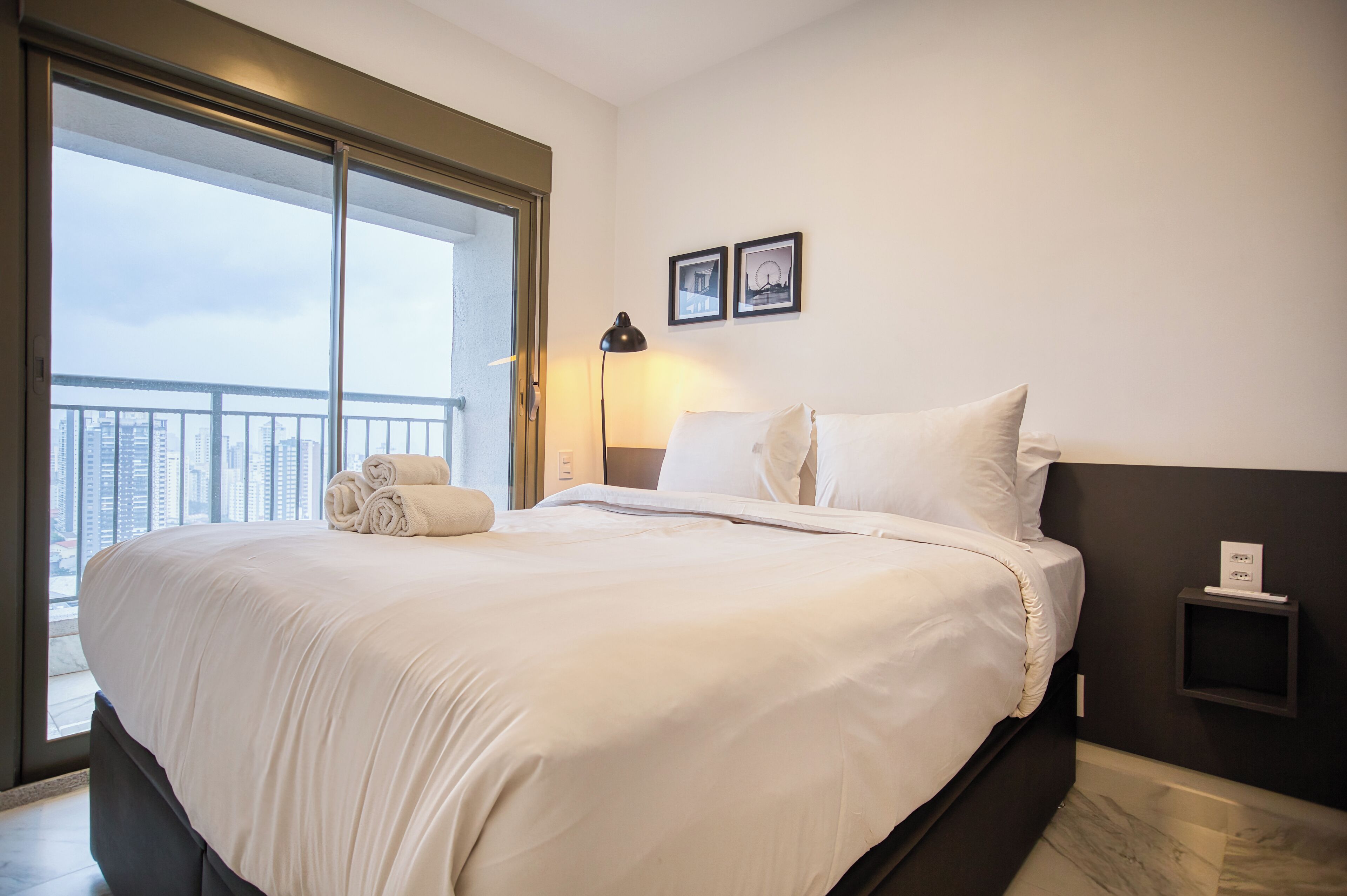 A hotel room with a large bed, a nightstand, a lamp, and a view of the city through a sliding door.