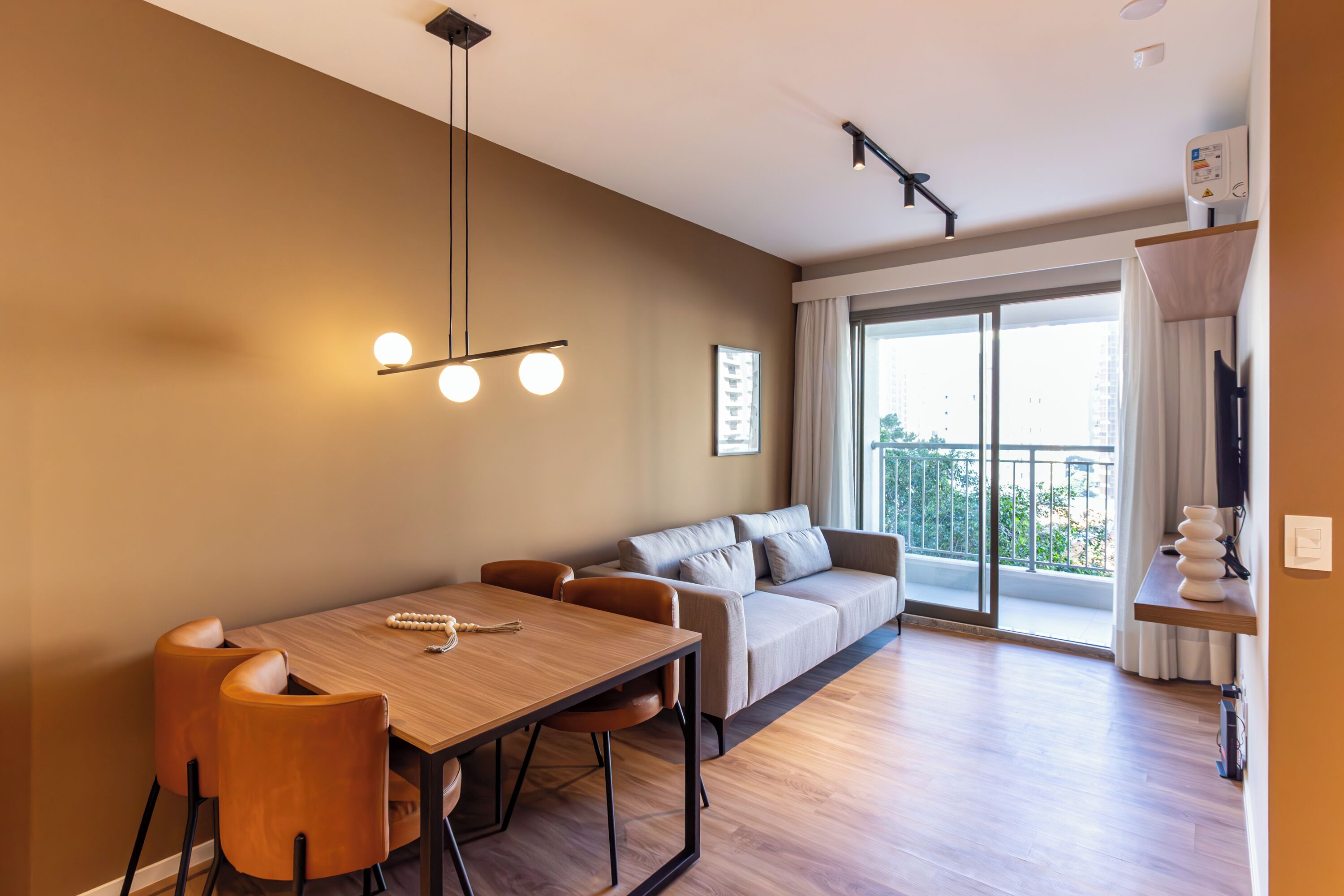 A modern living room with a wooden dining table, leather chairs, a sofa, and a large sliding glass door to a balcony.