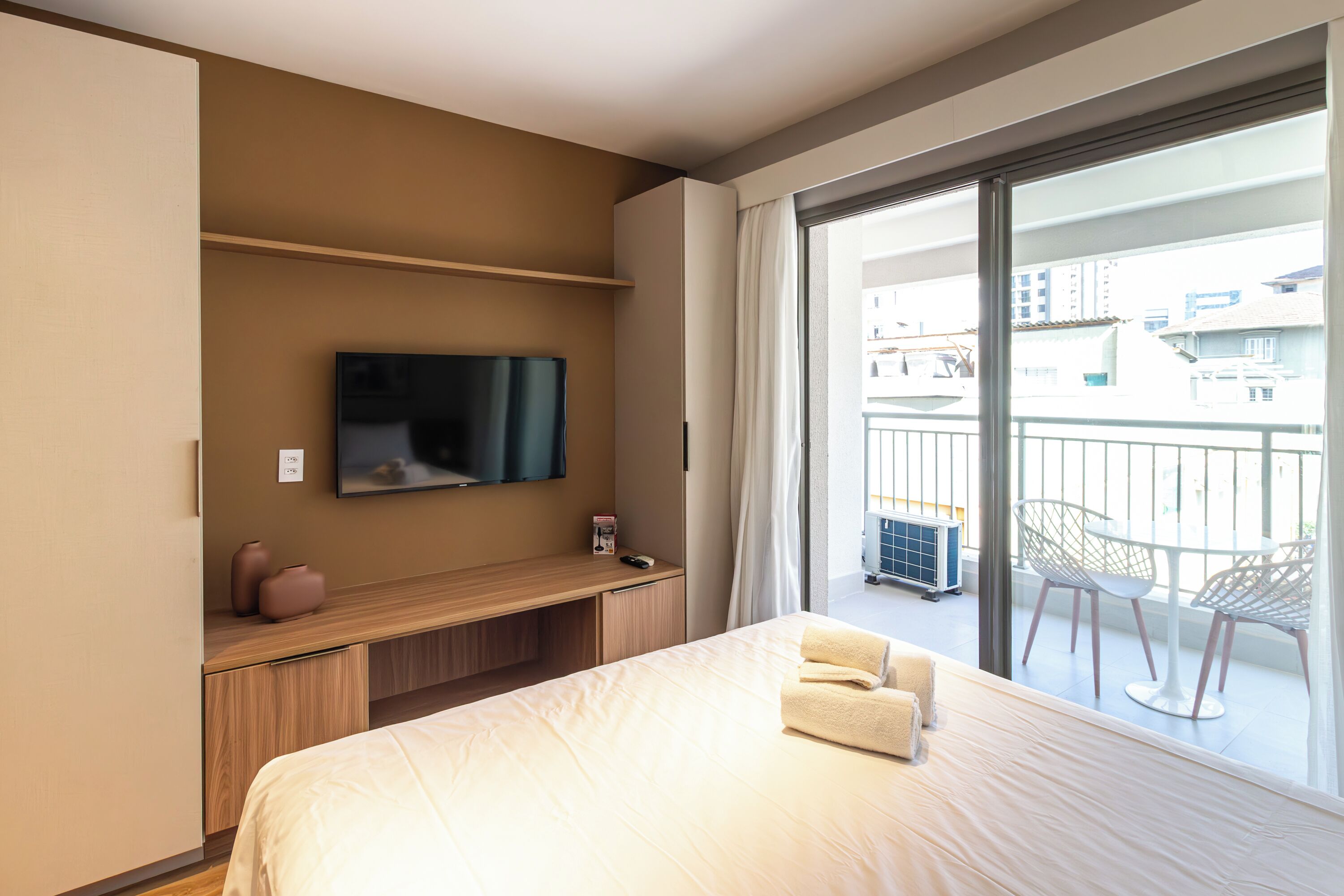 A modern hotel room with a large bed, a TV mounted on the wall, a wooden desk, and a balcony with white chairs.