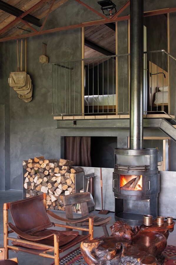 A rustic interior with a wood-burning stove, stacked firewood, a leather chair, and a wooden sculpture.