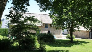 Een rustiek stenen gebouw met rieten dak, omgeven door weelderige begroeiing en bomen.