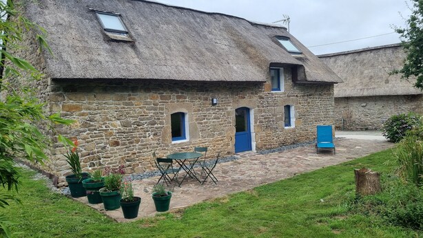 Een gebouw van steen met rieten dak, een klein terras met een tafel en stoelen, en potplanten.