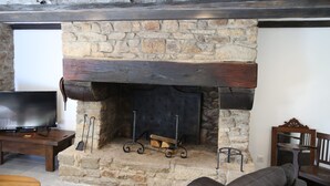 Een stenen openhaard met houten bovenblad, een televisie op een houten dressoir links, en een leren bank aan de rechterkant.