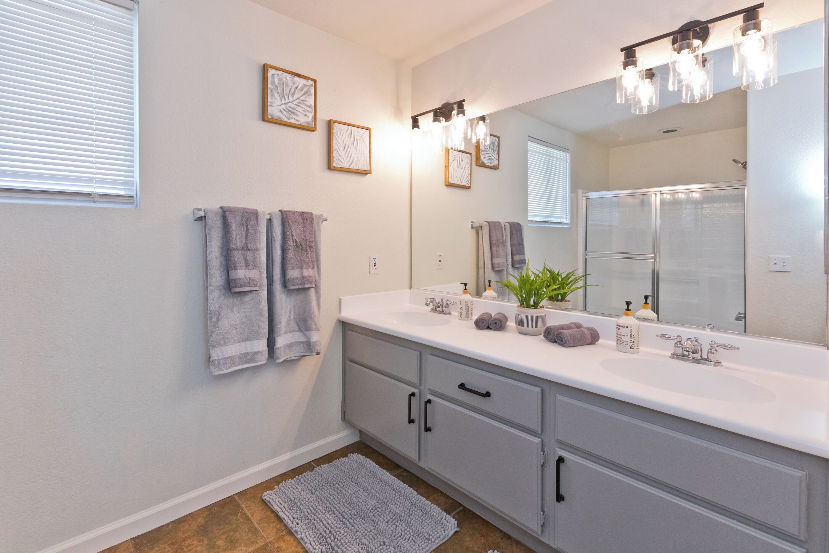 A bathroom with a large mirror, two sinks, and a shower. There are towels hanging on the wall and a small plant on the counter.