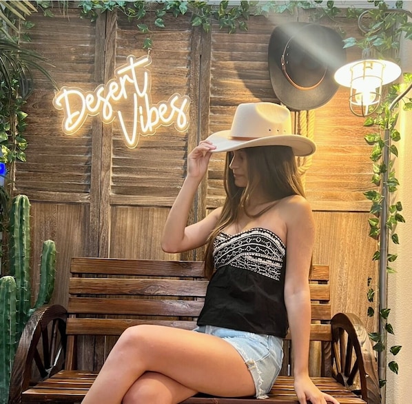 Una persona sentada en un banco de madera en un entorno rústico, con un letrero de neón que dice “Desert Vibes” y una lámpara antigua.
