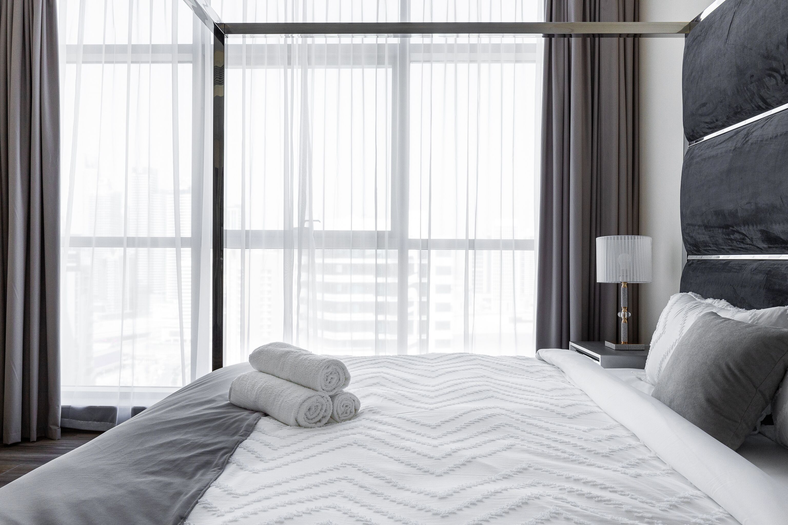 Habitación de hotel con una cama grande, una mesita de noche con lámpara y vistas a la ciudad a través de amplios ventanales.
