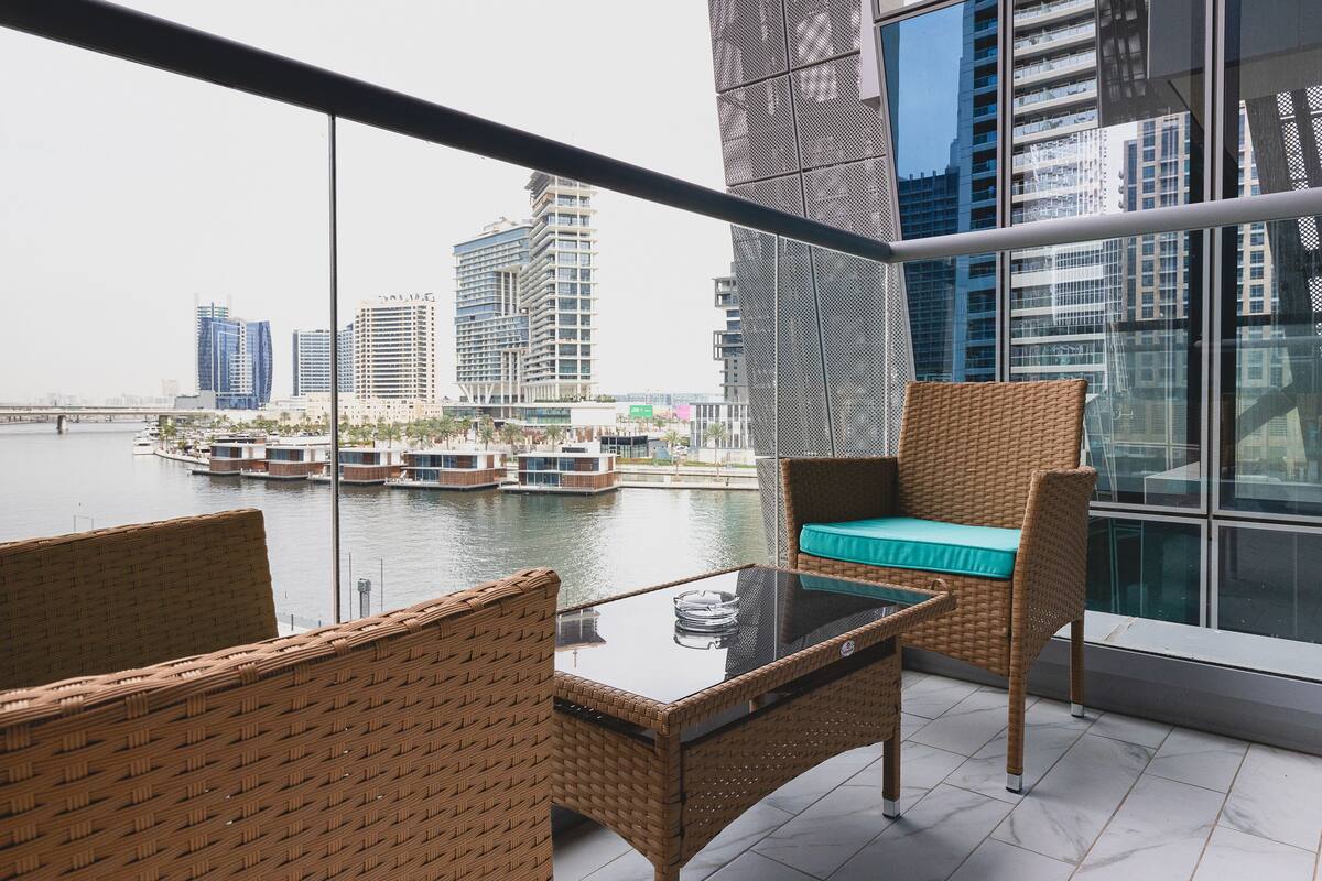 A balcony with wicker furniture, a glass table, and a view of a cityscape and water.