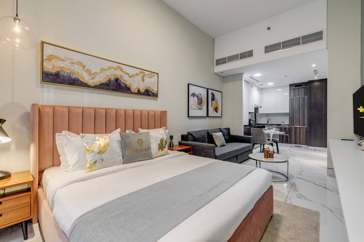 A modern hotel room with a large bed, a sofa, a small round table, and a kitchenette in the background.
