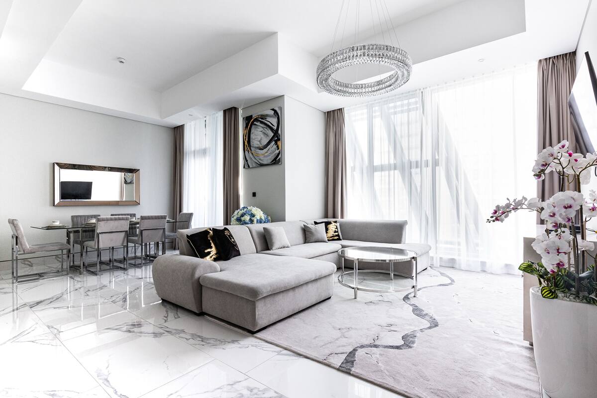 A modern living room with a sectional sofa, a dining area with a glass table, and a large window with sheer curtains.