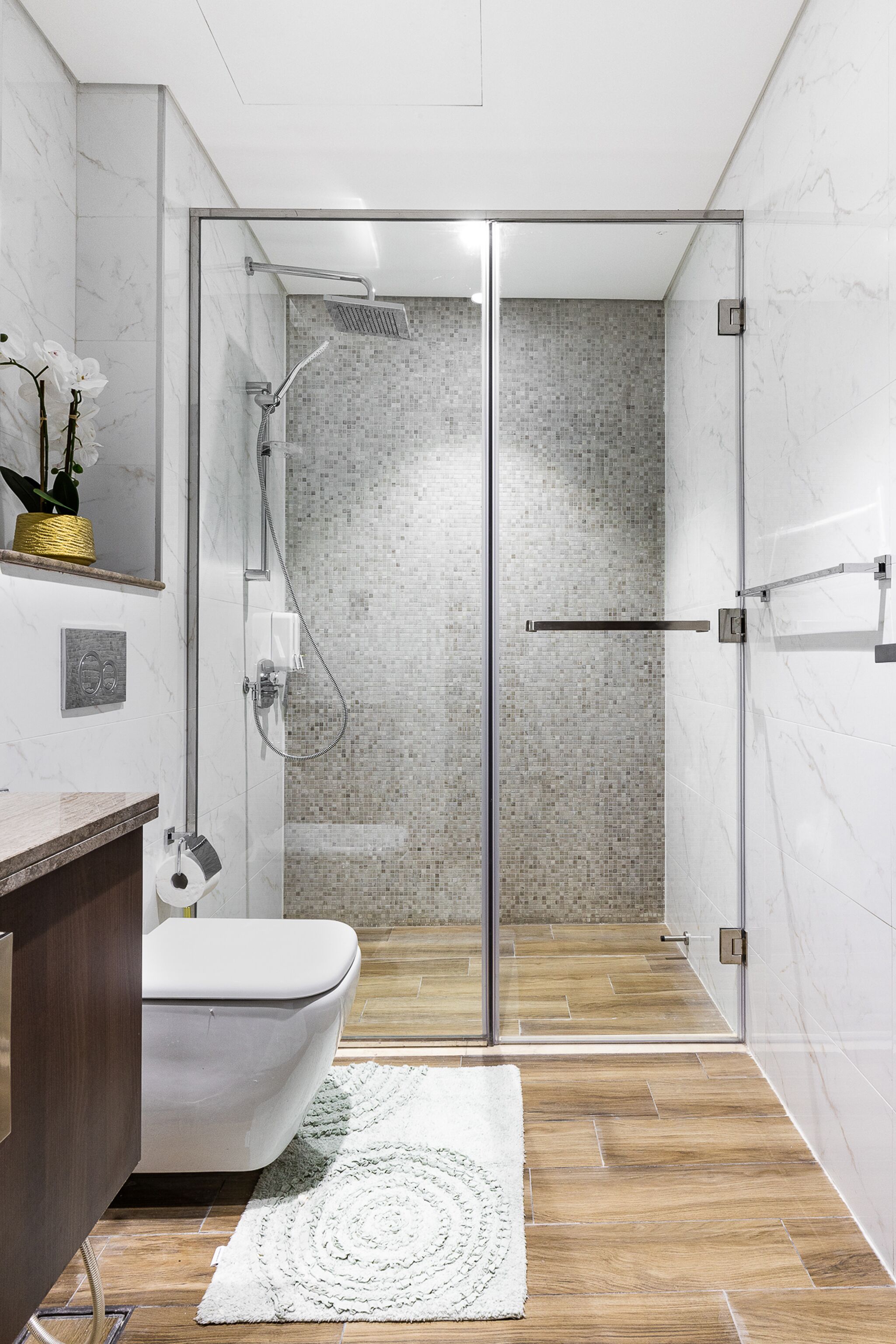 Ein modernes Badezimmer mit einer gläsernen Duschkabine, einer weißen Toilette, einem hölzernen Waschtisch und einem gemusterten Teppich.