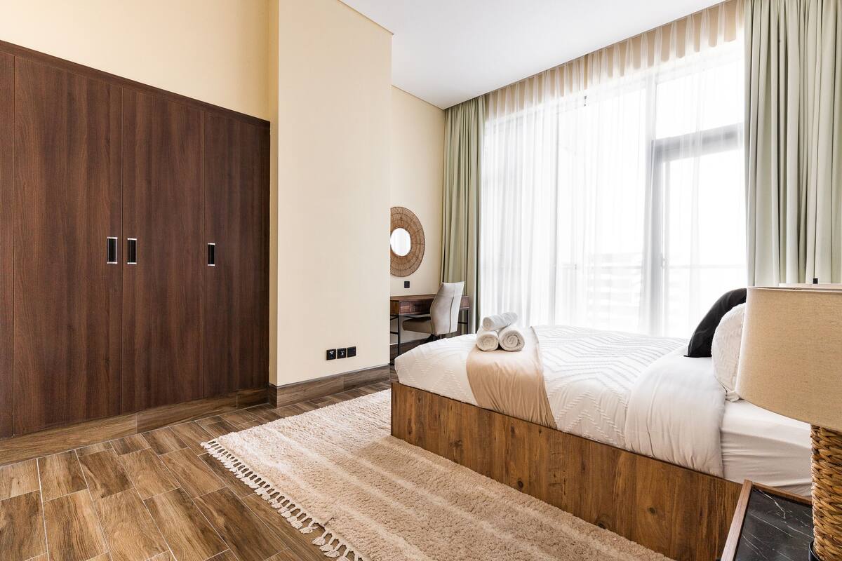A modern bedroom with a large wardrobe, a bed with a wooden headboard, a round mirror, and a view of the outside through sheer curtains.