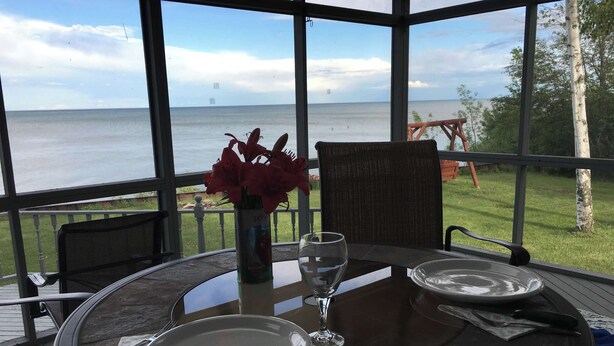 Une table avec deux assiettes et un verre à vin, un vase rempli de fleurs rouges et une vue sur la mer par de grandes fenêtres.