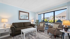 Una sala de estar con un sofá marrón, dos sillones estampados, una mesa de centro blanca y un ventanal con vistas a una zona residencial.