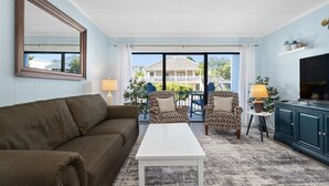 Una sala de estar con un sofá marrón, una mesa de centro blanca, sillones estampados, un televisor y vistas a una casa a través de amplios ventanales.