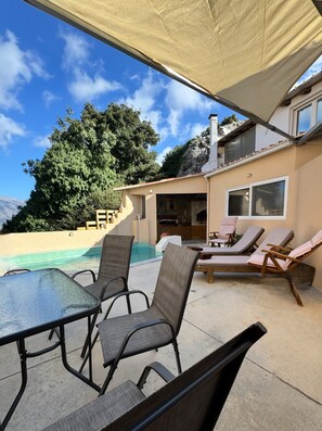 A poolside area with lounge chairs and a table, a covered patio, and a view of a mountain and trees.