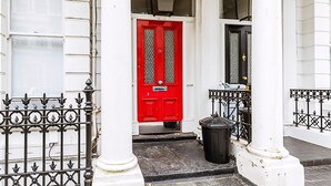 A red door with a number plate, white columns, and a black trash bin in front. - Lovely One-bedroom Flat Near West Kensington (London)
