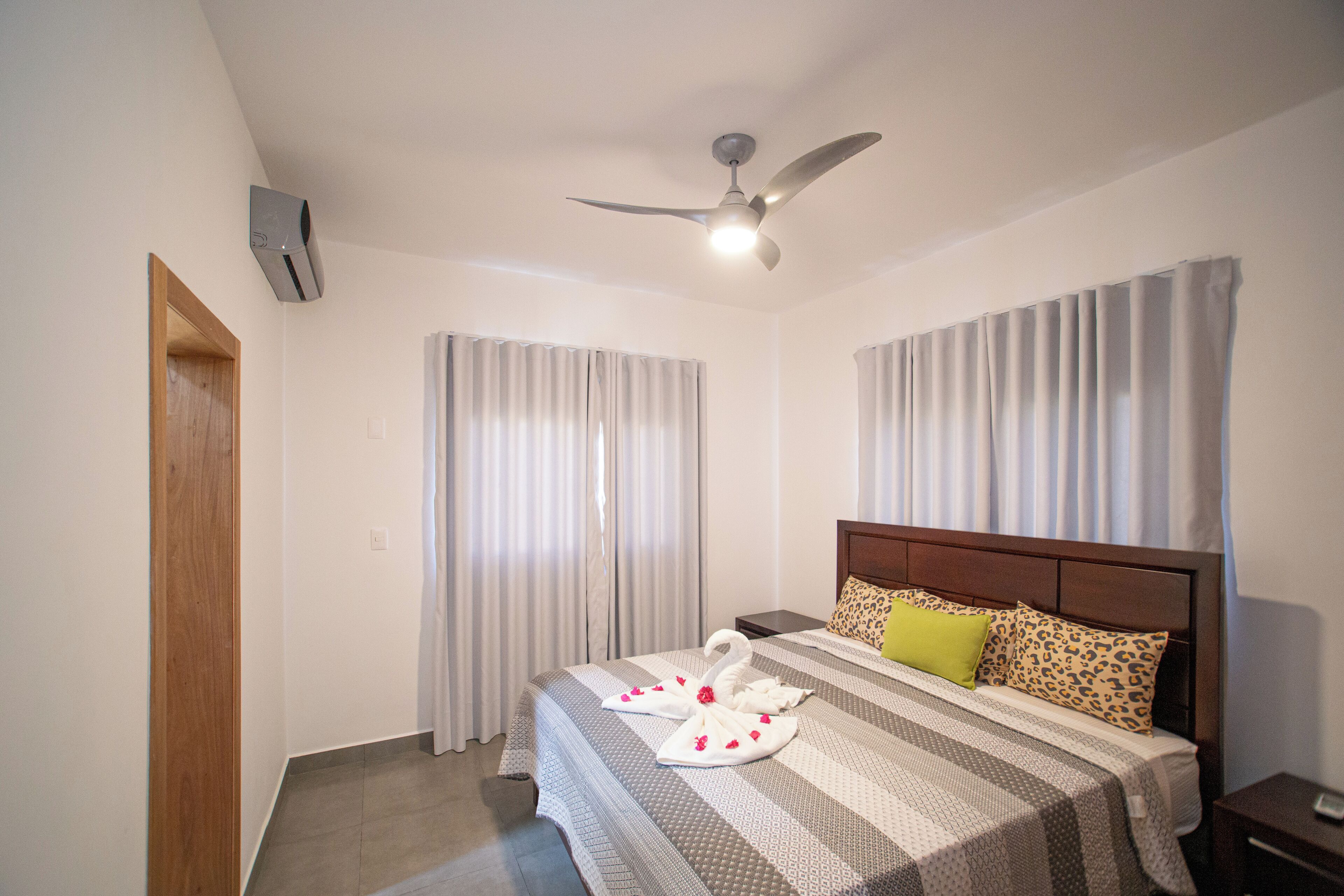 A hotel room with a bed, a ceiling fan, curtains, and a wooden door.