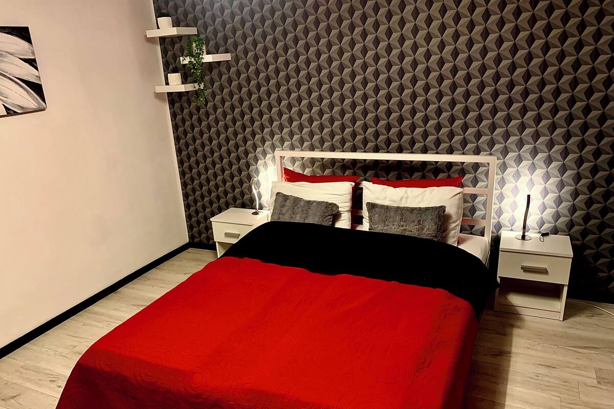 A bedroom with a bed, white nightstands, a red bedspread, and a geometric patterned wall.