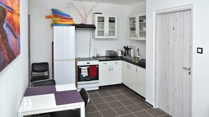 Cocina pequeña con gabinetes blancos, estufa y pileta. Se ve una mesa de comedor con sillas y un cuadro en la pared.