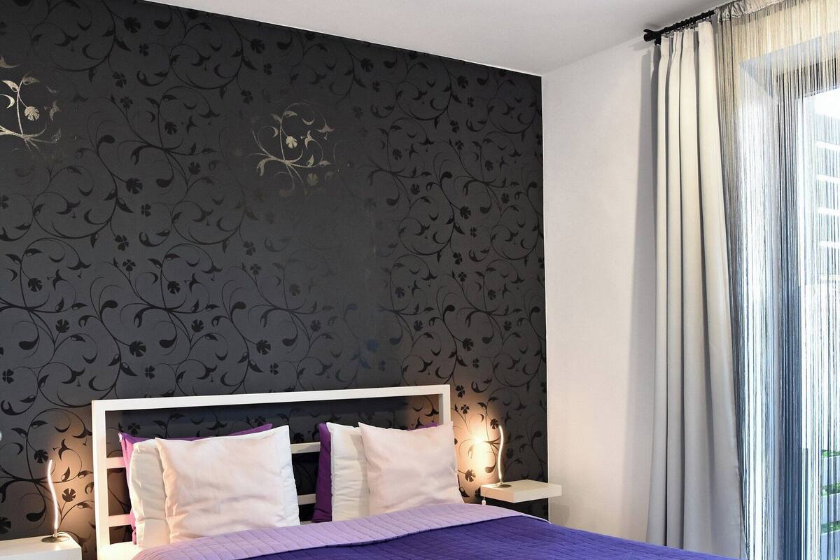 A modern bedroom with a large bed, a patterned wallpaper, and a view of the outdoors through a window with curtains.