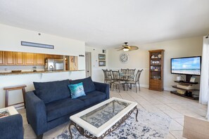 Un salon comprenant un canapé, une table basse et un téléviseur. On y trouve également un coin repas avec des chaises et une table. La cuisine est visible et on peut y apercevoir des placards et des appareils électroménagers.