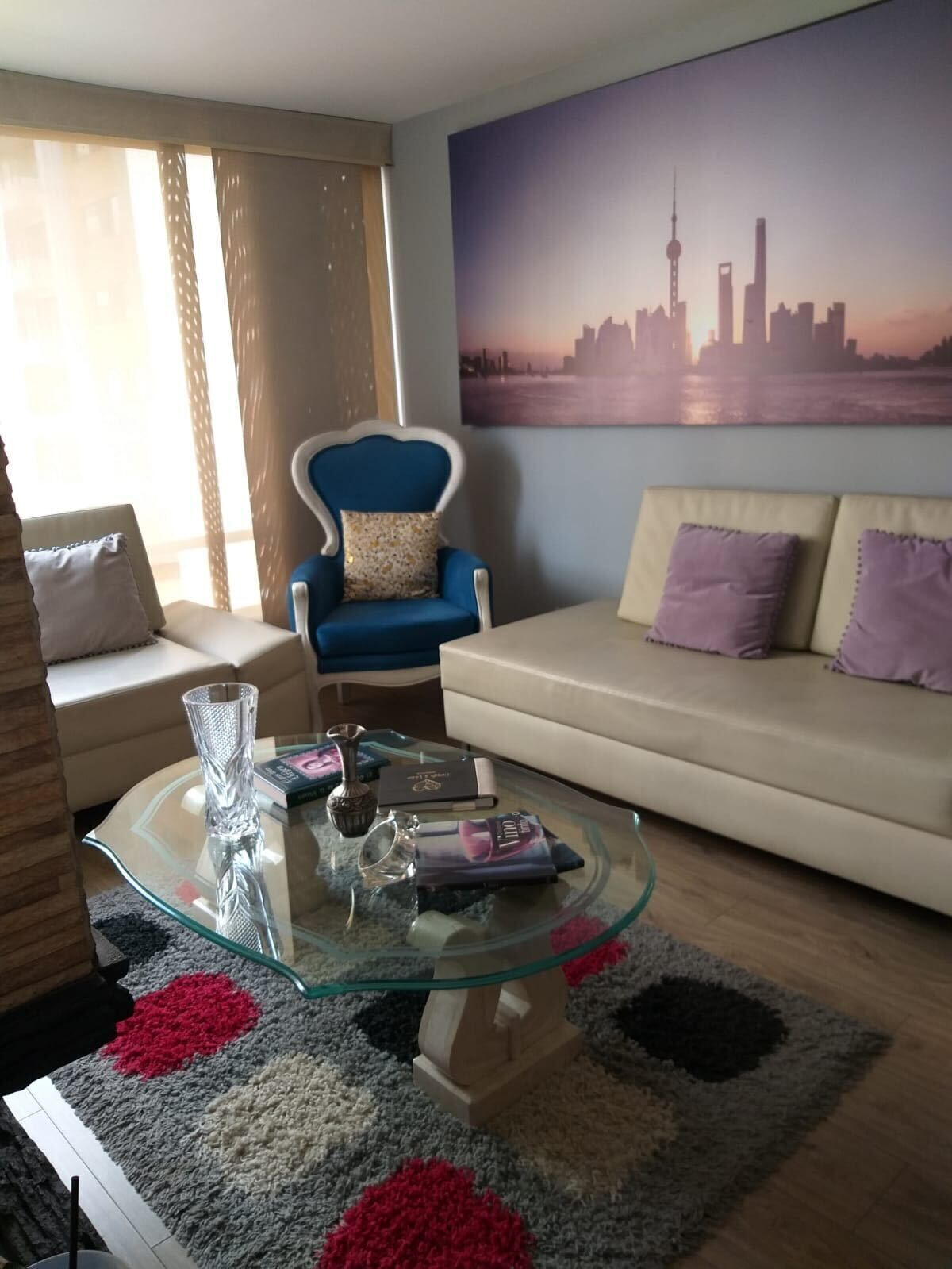 Een moderne woonkamer met een glazen salontafel, een blauwe fauteuil, een beige bank en een stadsgezicht schilderij aan de muur.
