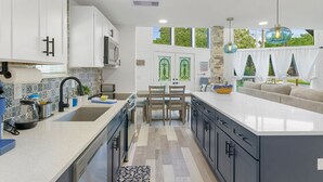 A modern kitchen with a central island, white cabinets, and a dining area with a table and chairs. - Private Coastal home w. gaming loft, pool table, hot tub, arcades - pet friendly (Hudson)