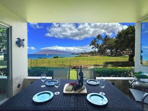 Une table à manger dressée avec des assiettes, des verres et une bouteille, offrant une vue panoramique sur une plage et des montagnes.