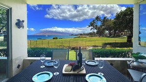 A dining table set with plates, glasses, and a bottle, overlooking a scenic view of a beach and mountains. - Discover Paradise at Royal Mauian | Ground Floor Oceanview and Island Living (Kihei)