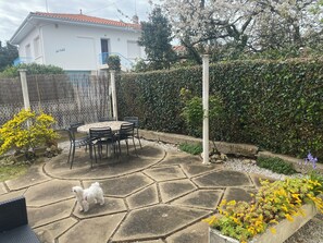 A patio with a stone table and chairs, surrounded by a hedge and flowering plants.