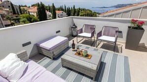 Une terrasse sur le toit, avec un canapé, deux chaises, une table avec une bouteille et des verres, et une vue sur la mer et les bâtiments.