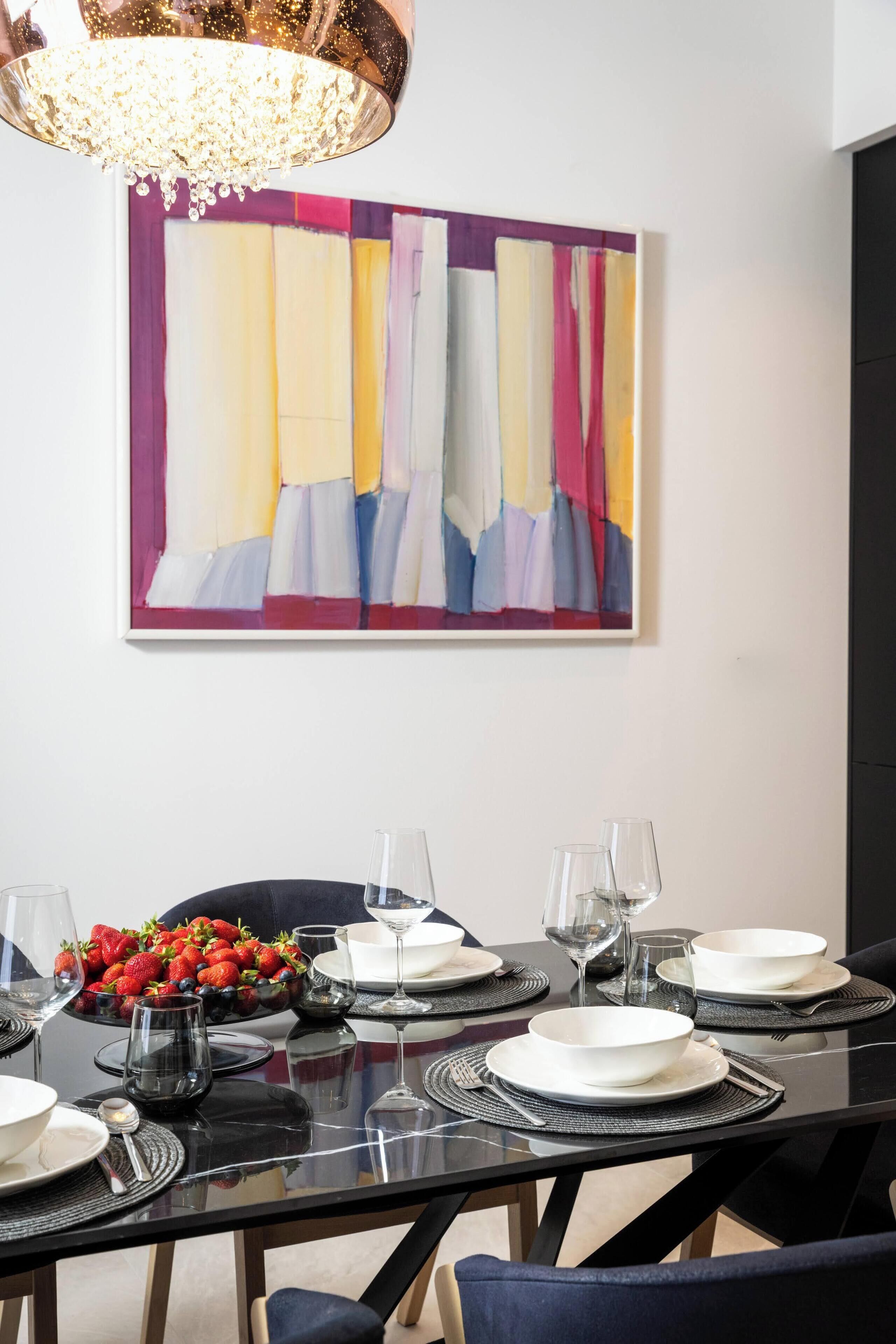 Un coin repas avec une table noire dressée pour quatre personnes, un tableau abstrait aux couleurs vives accroché au mur et un lustre au-dessus.