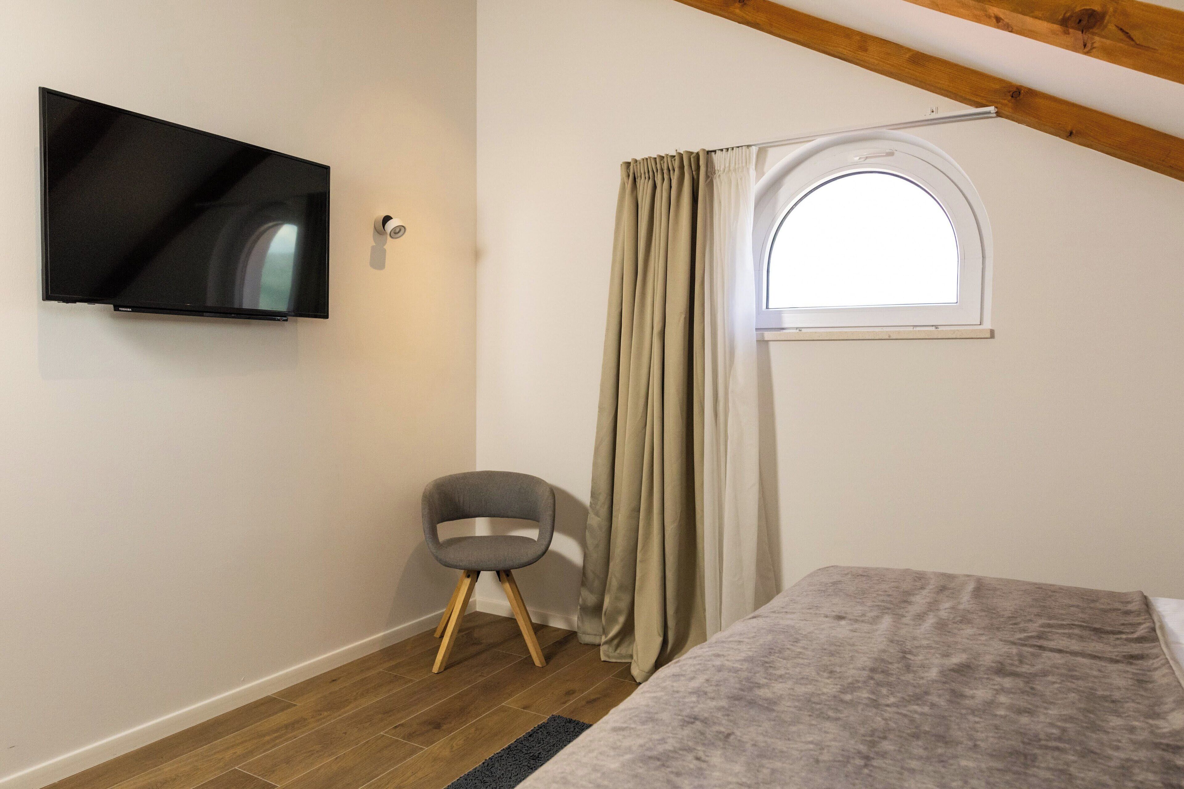 Una camera da letto con una TV fissata al muro, una finestra con tende, una piccola sedia e un letto con coperta.