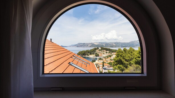 Une vue depuis une fenêtre à cadre incurvé, donnant sur une ville côtière aux toits de tuiles rouges, avec une végétation luxuriante et des montagnes à l’horizon.