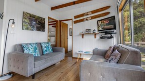 A spacious living room with a grey sofa, a smaller grey armchair, wooden beams on the ceiling, a television on a wooden shelf, and a large window with a view of trees.