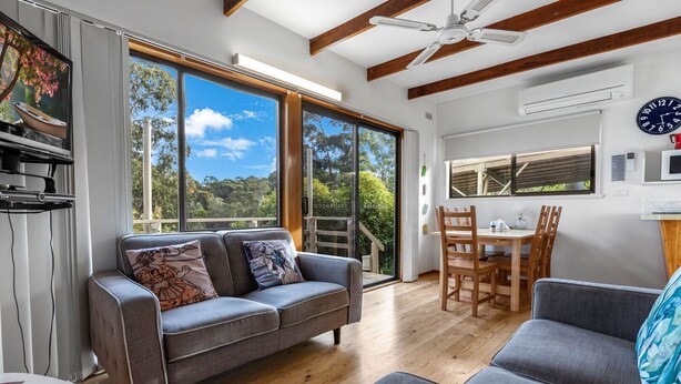 A modern living room with a grey sofa, wooden dining table, and large windows offering a view of the outdoors.