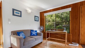 A room with a grey sofa, wooden flooring, a wooden desk, and a window with a view of greenery.