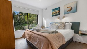 A bedroom with a large window, a bed with a beige bedspread, two wall-mounted lamps, and two paintings.