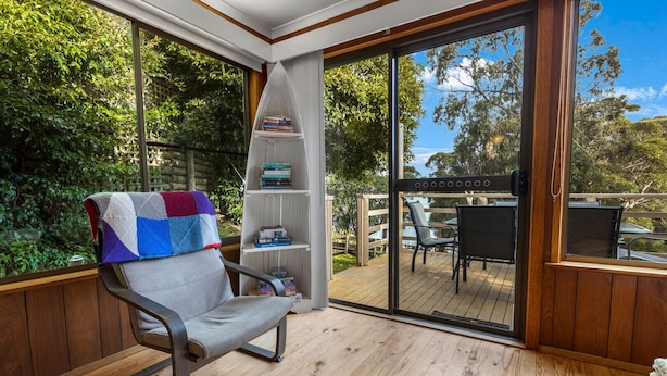 A cozy reading nook with a cushioned chair, a bookshelf, and a view of a wooden deck and greenery.