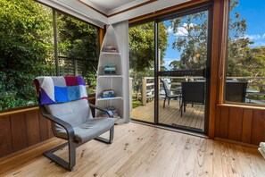A cozy reading nook with a cushioned chair, a bookshelf, and a view of a wooden deck and greenery. - Jetty View - Pelican Cabin: Feel a sense of nature in this rural environment (Nungurner)