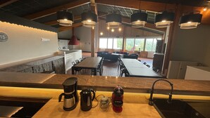 Una cocina moderna con una encimera larga, una cafetera, un hervidor y un hervidor eléctrico rojo. En la zona de comedor hay varias sillas y mesas, y ventanas amplias con vistas a árboles.