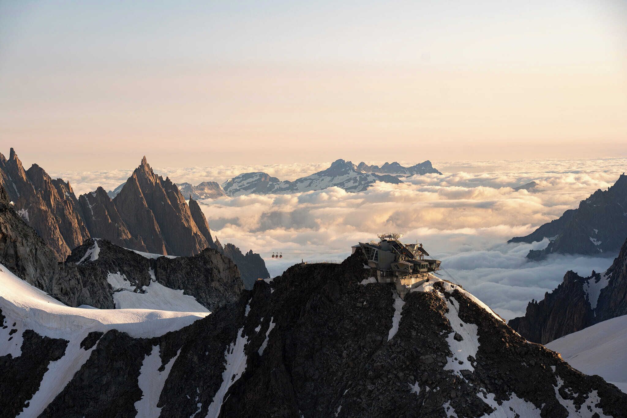 A mountain peak with a small structure on top, surrounded by snow-covered terrain and a layer of clouds below.