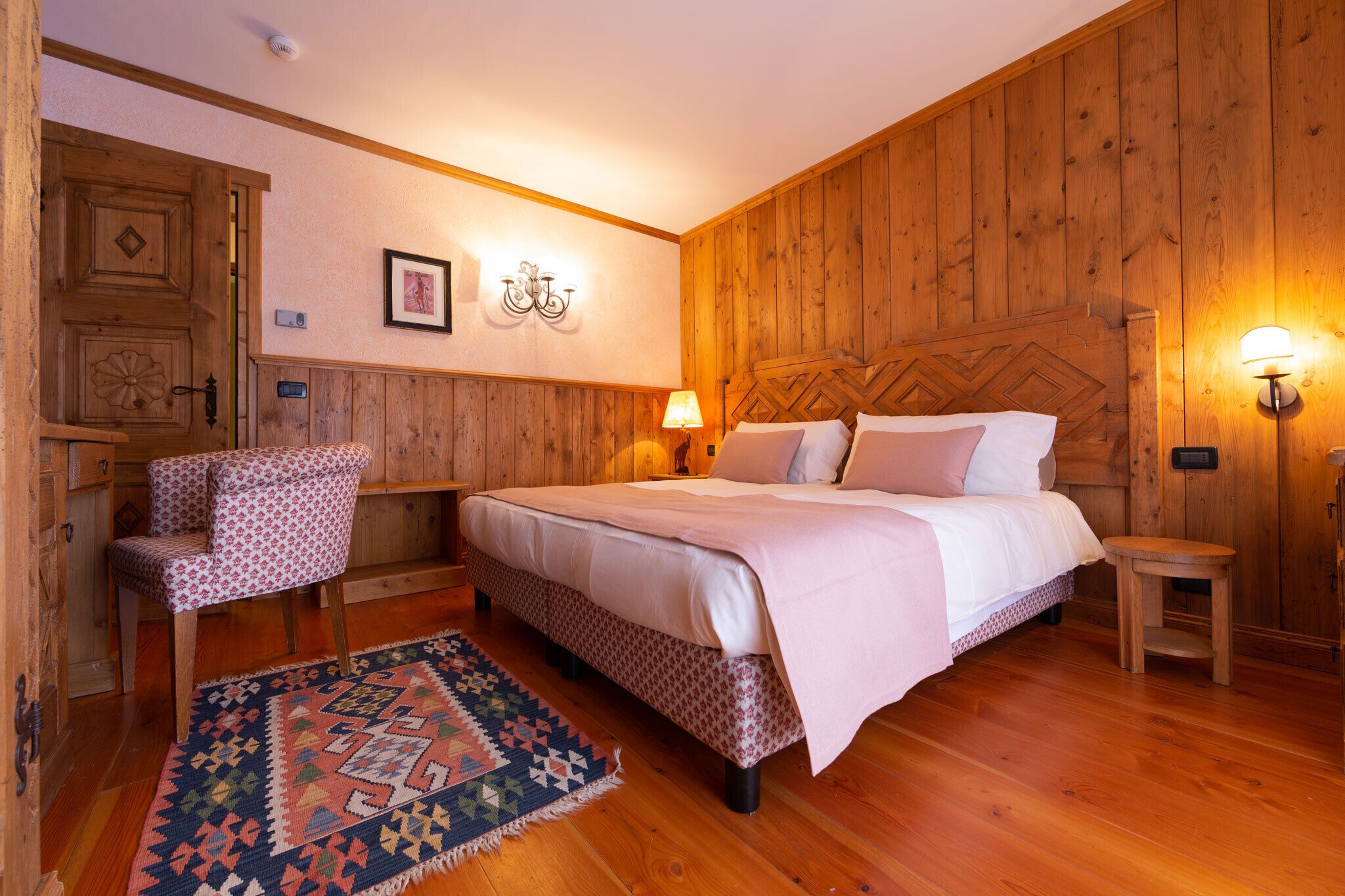 A wooden-paneled bedroom with a bed, a chair, a rug, and wall-mounted lamps.